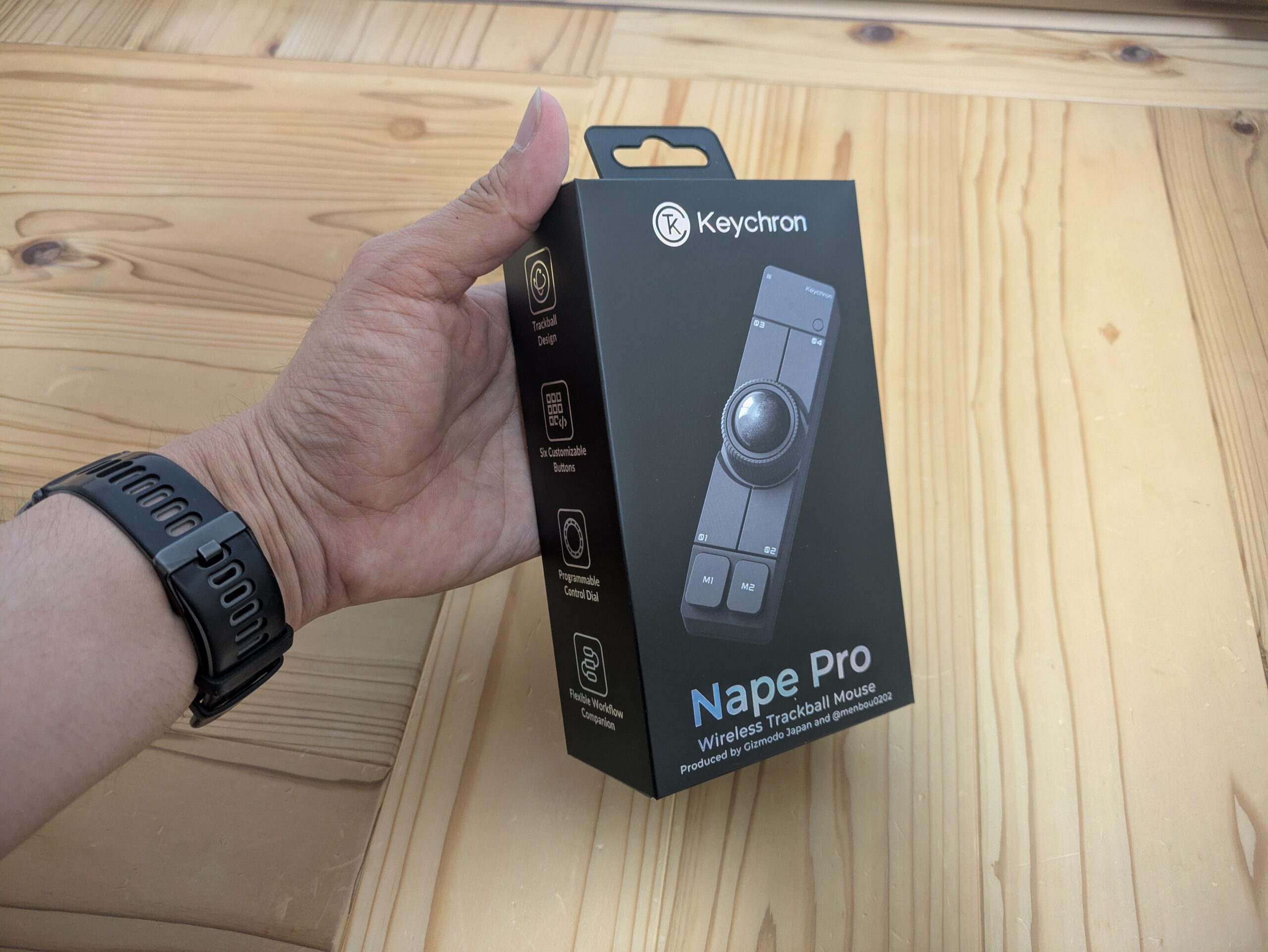 木の机の上で、手に持ったKeychron Nape Proの外箱を撮影した実写写真。黒いパッケージには、ワイヤレストラックボールマウス本体の画像と「Nape Pro」「Wireless Trackball Mouse」の文字があり、トラックボールデザイン、6つのカスタマイズボタン、プログラム可能なコントロールダイヤルなどの特徴が表示されている。Keychron Nape Proの購入・レビュー・開封前の様子を伝える写真。