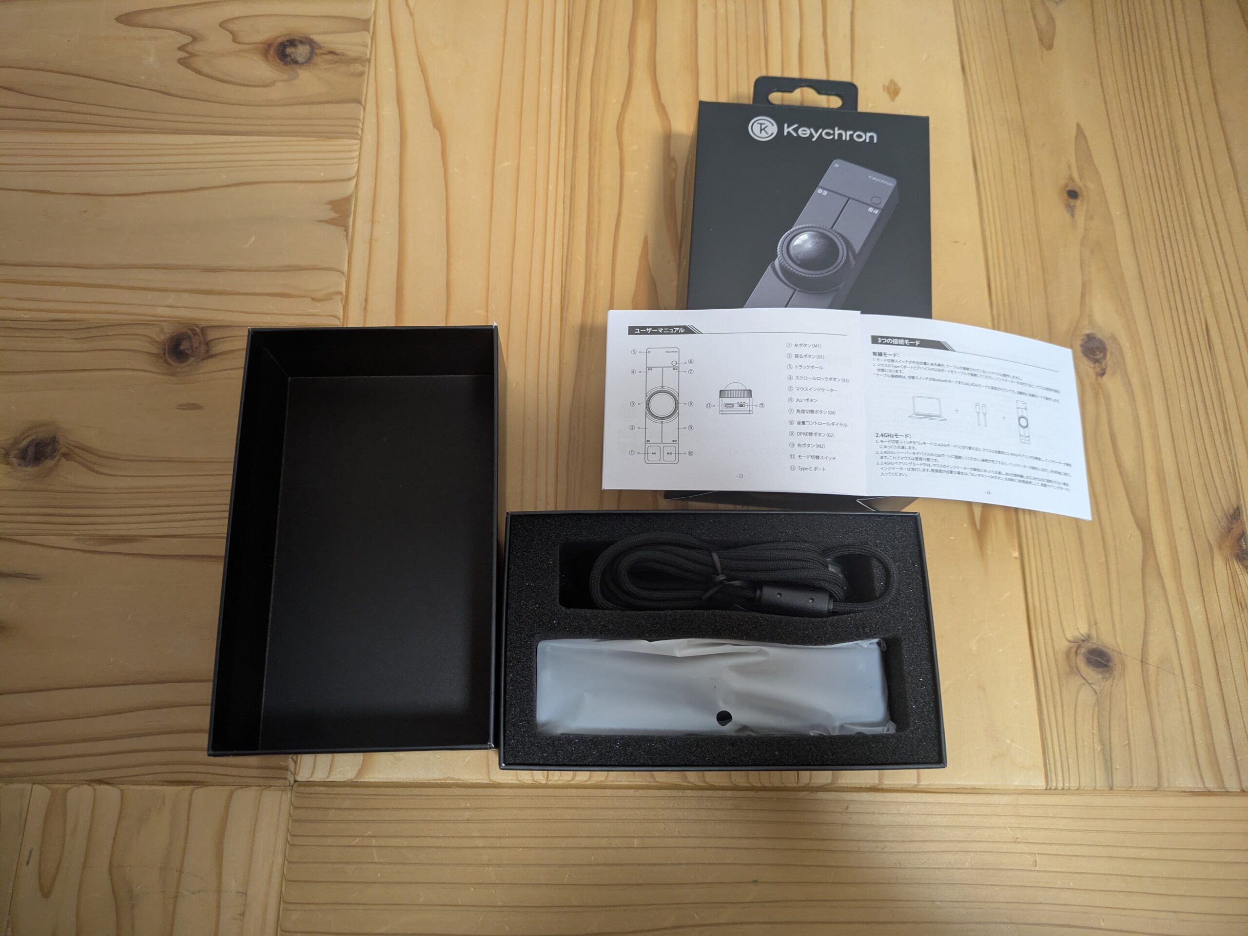 Keychron Nape Proを開封し、外箱、説明書、本体、USB-Cケーブルを木の机の上に並べた写真。箱の中には保護袋に入った本体と付属ケーブルが収納されており、開封時の同梱内容が確認できる。