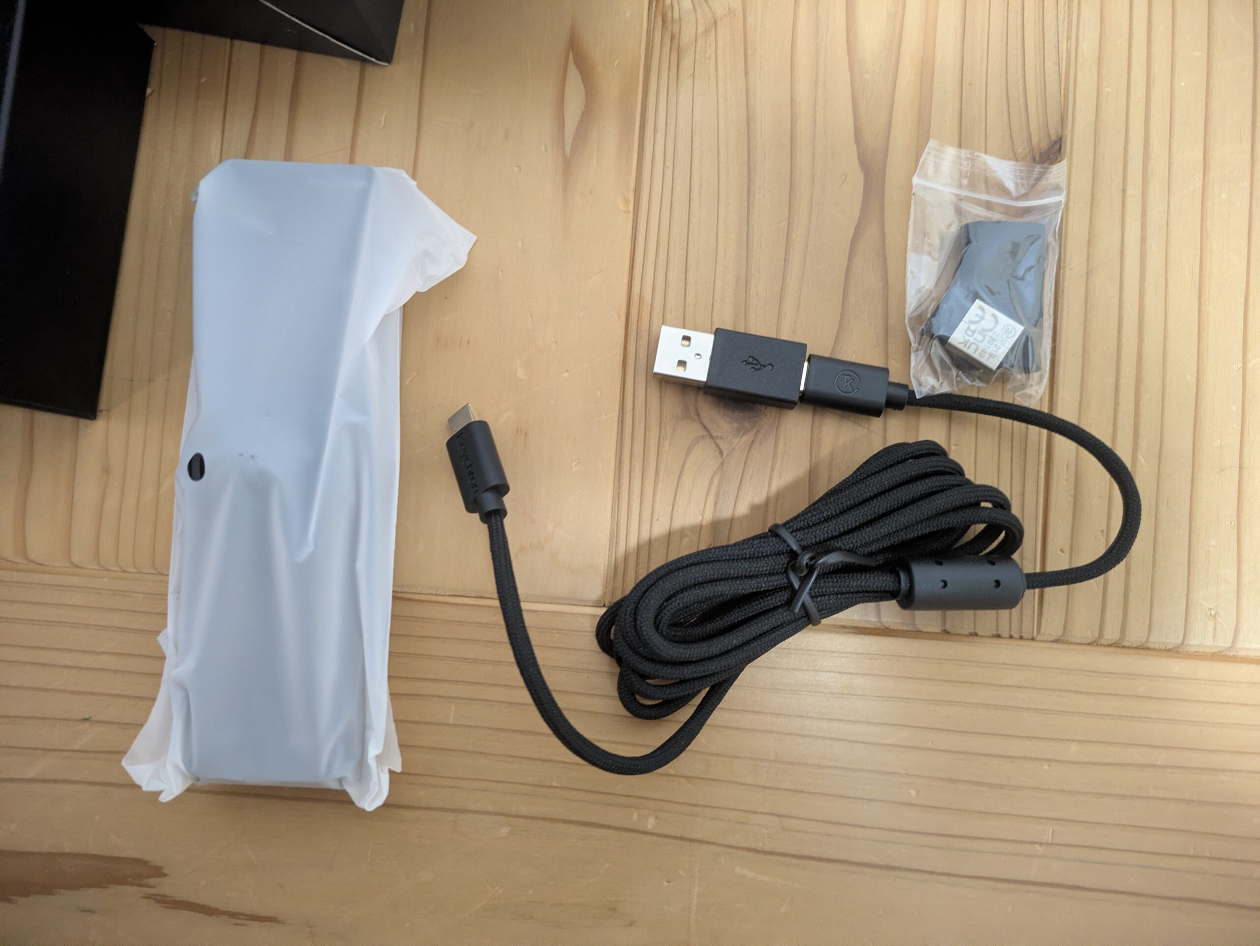 Keychron Nape Proの付属品を木の机の上に並べた写真。保護袋に入った本体、USB-Cケーブル、USBレシーバー、交換用パーツのような小物が確認できる。