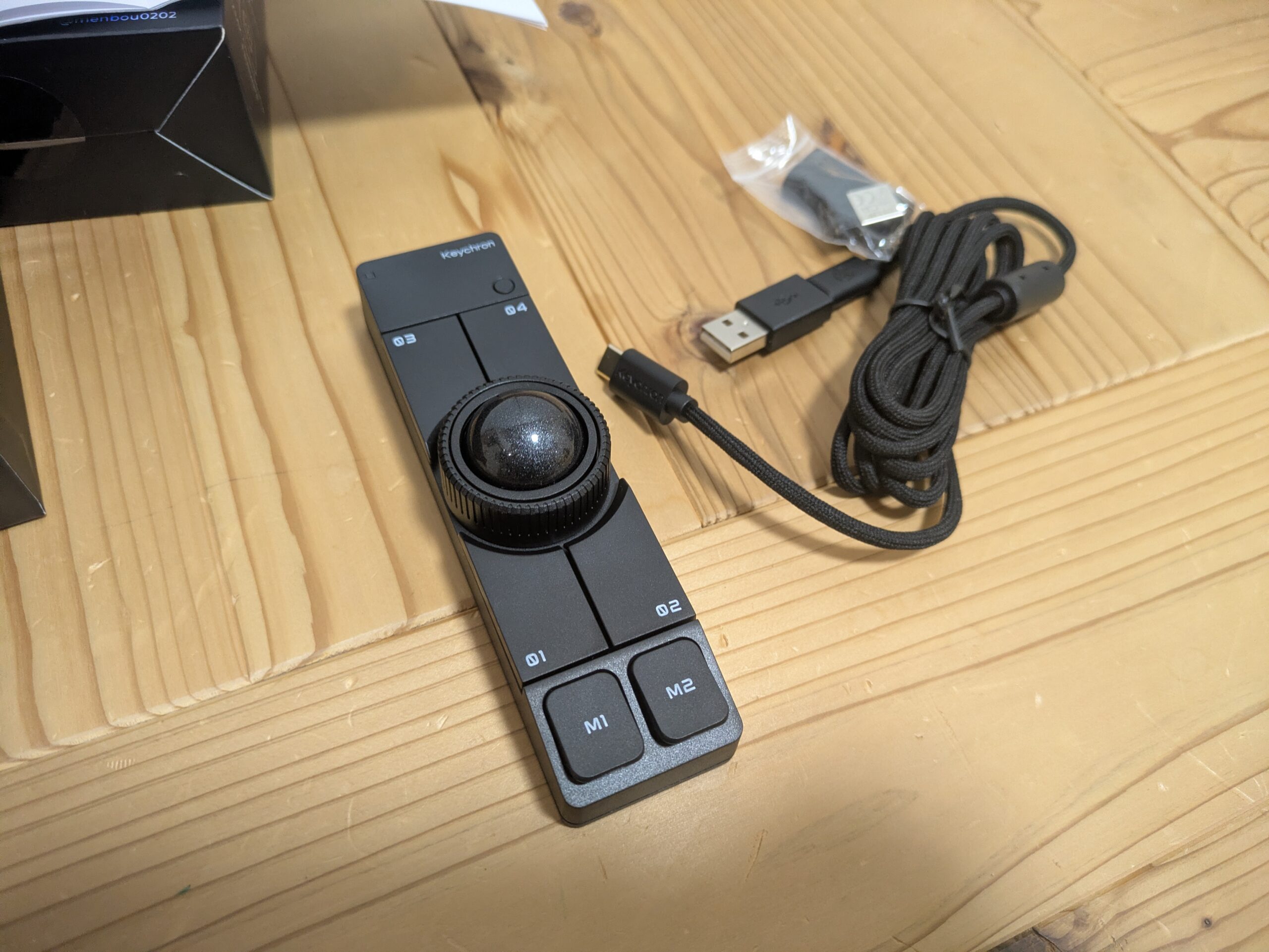 木の机の上に置いたKeychron Nape Pro本体と付属品の写真。中央にトラックボールを備えた細長い黒い本体の横に、USB-Cケーブル、USBレシーバー、交換用パーツが並んでいる。