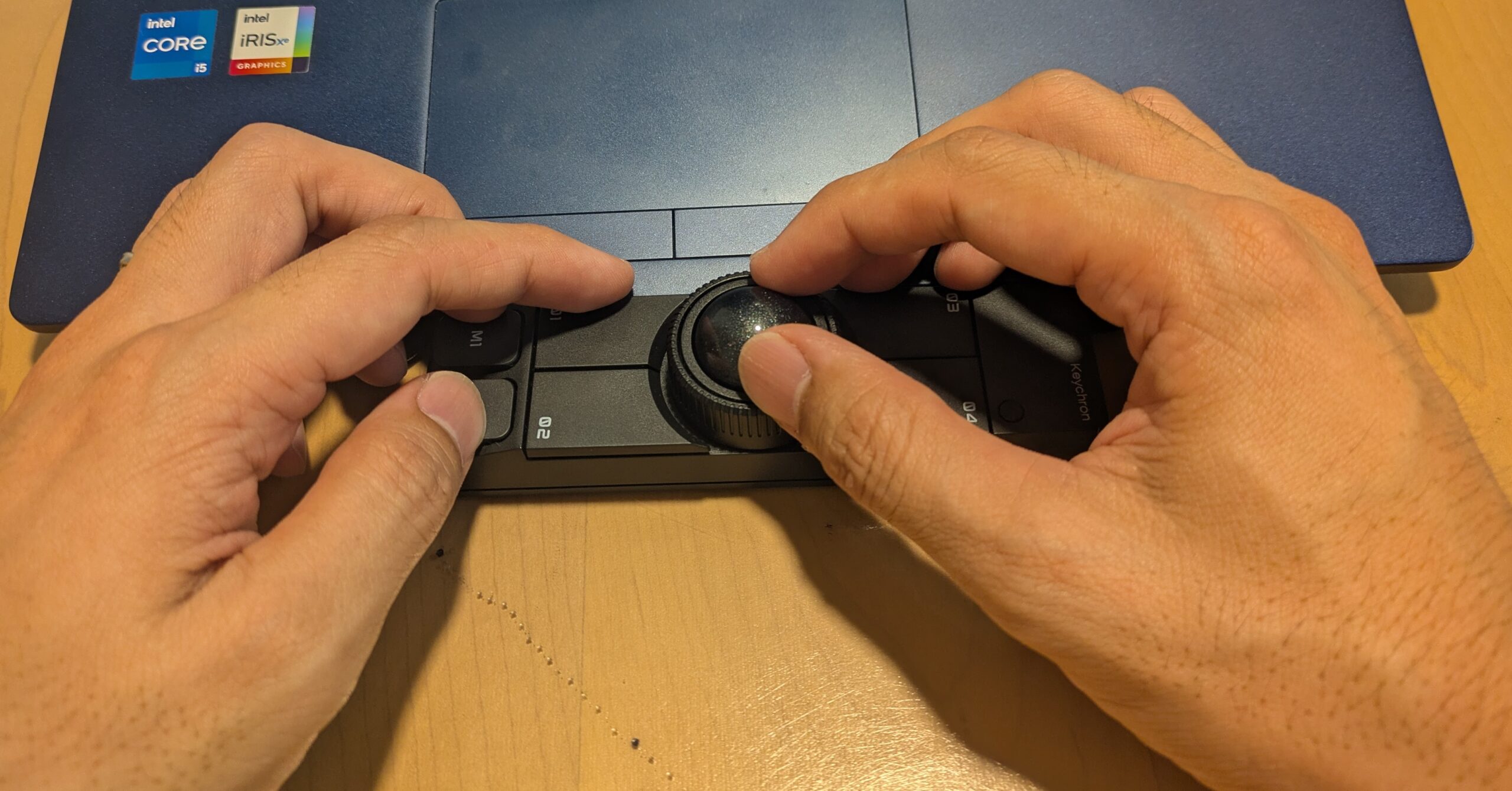 ノートパソコンの手前に置いたKeychron Nape Proを両手で操作している写真。右手の親指で中央のトラックボールを動かし、左手でM1・M2ボタン付近を押しており、実際の両手操作やショートカット操作の使い方を確認できる。