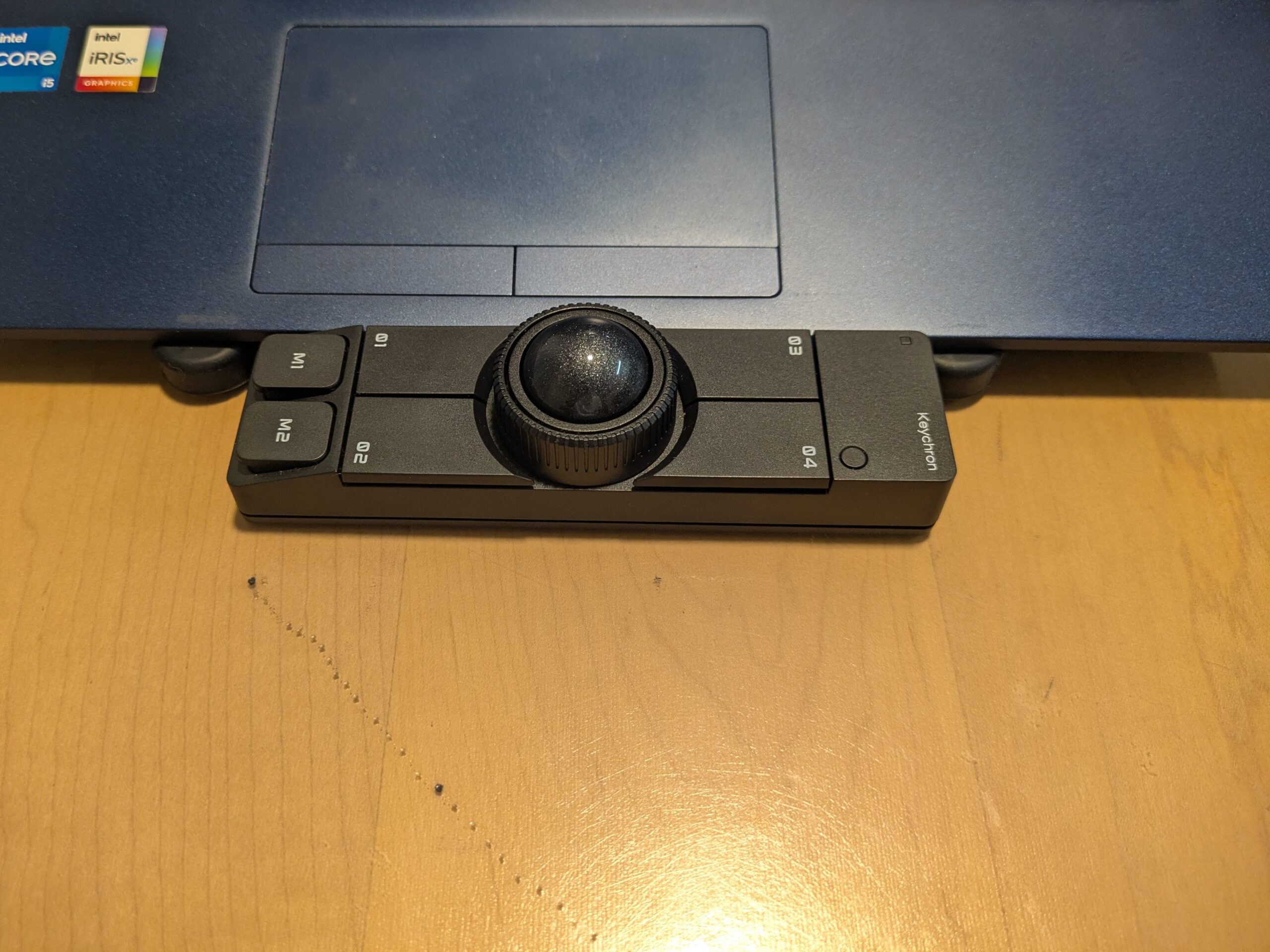 ノートパソコンの手前にKeychron Nape Proを置いた写真。細長い黒い本体の中央にトラックボールがあり、M1・M2ボタンやQ1〜Q4のカスタムボタンが見える。省スペースでノートPC前に置いたときのサイズ感や配置を確認できる。